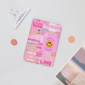 May include: A pink phone case with a collage of inspirational words and phrases, including "God is Good" and "Jesus Saves." The case features a smiley face and a cross. Other items include a white clip, two orange circles, and a newspaper.