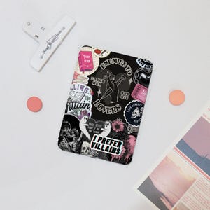 Peut inclure: Une coque de téléphone noire ornée d'autocollants sur les thèmes gothique et romantique. Les autocollants incluent des phrases comme « Enemies to Lovers » et « I Prefer Villains », ainsi que des illustrations de crânes, de fantômes et de fleurs. Une pince blanche et deux cercles roses sont également dans l'image.