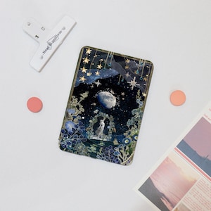 Könnte beinhalten: Ein rechteckiges Harz-Kunstwerk mit einem dunkelblauen Hintergrund, das einen Halbmond, Sterne und eine Figur zeigt, die in einem floralen Torbogen steht. Das Werk hat einen goldenen Rahmen und ist mit goldenem Glitzer und blauen und goldenen Blumen verziert.