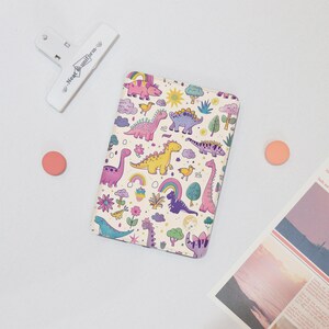 May include: A white notebook cover featuring a colourful dinosaur pattern. The dinosaurs are pink, purple, yellow, green, and blue. The background is white with clouds, rainbows, and flowers.
