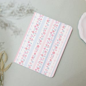 May include: A floral patterned notebook with a pink, blue, and white design. The notebook features rows of pink flowers, blue gingham, and dotted lines with bows. The cover has rounded corners.