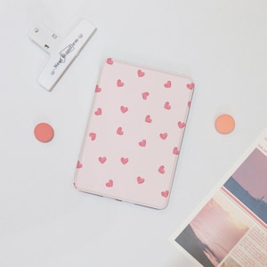 Può includere: Una custodia per tablet rosa chiaro con un motivo di piccoli cuori rossi. Una clip bianca con la scritta "Near Uniform" è in alto a sinistra. Due piccoli cerchi arancioni e un giornale sono anche nell'immagine.