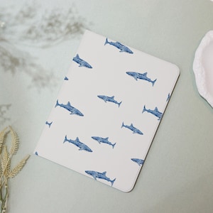 May include: A white notebook with a pattern of blue sharks. The sharks are stylized and evenly spaced across the cover. The notebook has rounded corners and a clean, minimalist design.