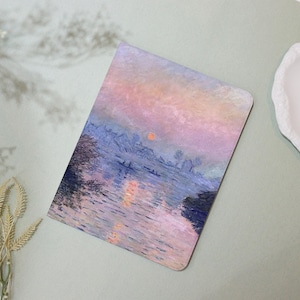 Könnte beinhalten: Ein rechteckiges Notizbuch mit einem pastellfarbenen Landschaftsgemälde. Das Kunstwerk zeigt eine Flussszene mit Sonnenuntergang in Rosa-, Blau- und Orangetönen. Das Design des Notizbuchs erinnert an den Impressionismus.