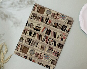 May include: A beige notebook cover with a repeating pattern of books on a bookshelf. The books are in various colors and sizes, and some have titles visible. The pattern is whimsical and charming.