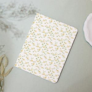 May include: A rectangular notebook with a white background covered in a pattern of small yellow roses and green leaves. The notebook is lying on a light green surface.
