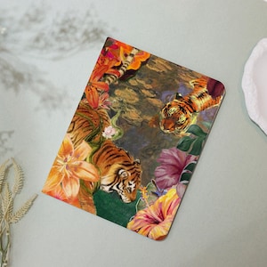 May include: A rectangular notebook featuring a vibrant illustration of tigers amidst lush, colorful flowers and foliage. The artwork showcases orange lilies, pink hibiscus, and a serene water scene, creating a tropical theme.