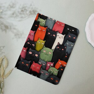 May include: A rectangular, black surface covered with a colorful illustration of numerous cartoon cats. The cats are in various colors, including black, red, green, and gray, with expressive faces and large eyes. The overall design is whimsical and playful.