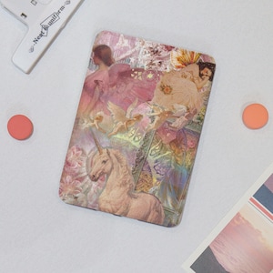 Peut inclure: Une housse de liseuse rectangulaire présentant un collage d'images roses et dorées, incluant une licorne, des figures ailées et des motifs floraux. Le design intègre des illustrations superposées avec une esthétique douce et une finition brillante.