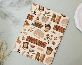 May include: A cream-colored notebook with a repeating pattern of books, lanterns, sleeping cats, and the word "BOOKSHOP". Other elements include keys, teacups, and potted plants. The notebook has a rectangular shape.