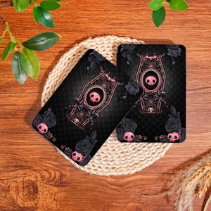 May include: Two black rectangular coasters with a pink skull coffin design, surrounded by black roses and pink skulls. The coasters are on a woven mat, with a wooden background.