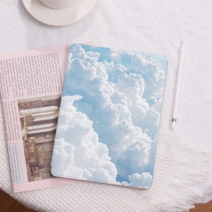 May include: A tablet case featuring a vibrant sky and cloud design in shades of blue and white. The case is rectangular and lies on a pink newspaper and a white, textured surface. A white stylus is also present.