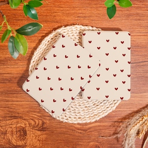 Puede incluir: Tres objetos rectangulares color crema con un patrón de pequeños corazones rojos. Los objetos están dispuestos sobre un salvamanteles circular tejido, sobre un fondo de madera. Se ve vegetación decorativa.