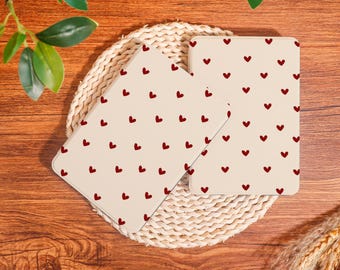 May include: Three cream-coloured rectangular objects patterned with small red hearts. They are arranged on a woven, circular placemat, set against a wooden background. Decorative greenery is visible.