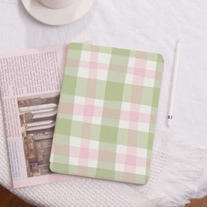 Pode incluir: Uma capa para iPad xadrez verde e rosa sobre fundo branco. A capa está sobre uma superfície branca com uma caneta branca ao lado.