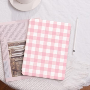 Op de afbeelding: Een tablet hoesje met een roze en wit ruitpatroon. De hoes is open en toont een krant. Een witte stylus en een witte theekop staan ook in de afbeelding. De hoes ligt op een wit, getextureerd oppervlak.