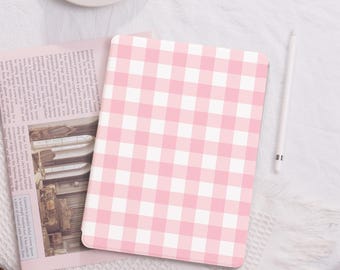 Può includere: Una custodia per tablet con motivo a quadretti rosa e bianchi. La custodia è aperta, rivelando un giornale. Nello scatto sono presenti anche uno stilo bianco e una tazza da tè bianca. La custodia è su una superficie bianca strutturata.