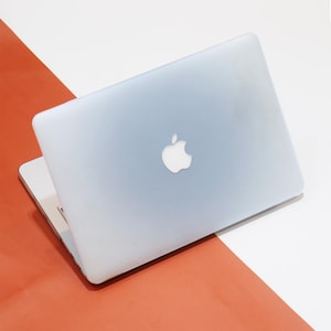 May include: A silver laptop with the Apple logo on the lid, slightly open. The laptop is set against a backdrop of white and orange surfaces. The laptop's screen is not visible.