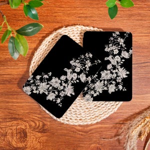 Pode incluir: Dois porta-copos retangulares com fundo preto e um padrão floral branco com ilustrações botânicas detalhadas de várias flores e folhas dispostas numa composição fluida e diagonal na superfície de cada peça.