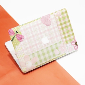 May include: A decorative laptop case featuring a patchwork design with pink and green floral and plaid patterns. The case includes a pink dragonfly, hearts, and flowers. The Apple logo is visible in the center.