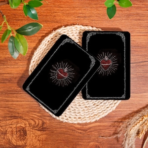 Puede incluir: Dos cartas de tarot negras con un diseño de corazón rojo, una corona y líneas radiantes. Las cartas están sobre un posavasos tejido, sobre un fondo de madera. El diseño del corazón incluye una cadena.