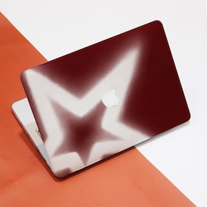 May include: A burgundy laptop with a white star design. The star is airbrushed onto the laptop's cover, creating a gradient effect. The Apple logo is visible on the laptop's surface. The laptop is open, revealing the keyboard.