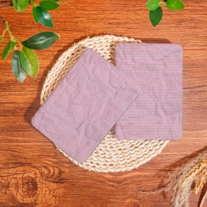 May include: Two pink lined paper sheets with grey lines rest on a woven straw mat, set against a wooden background. The paper has a slightly crumpled texture. Green leaves and wheat stalks are visible.
