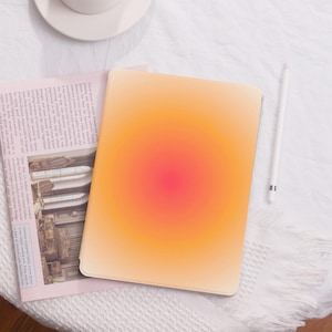 Könnte beinhalten: Nahaufnahme eines Tablets mit einem Farbverlaufshintergrund, der von einem hellen Orange zu einem leuchtenden Pink in der Mitte übergeht. Das Tablet liegt auf einer weißen Oberfläche mit einem weißen Stift daneben.