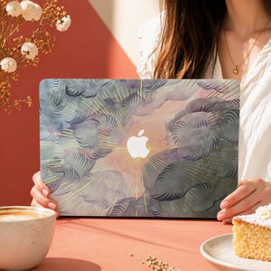 Può includere: Una skin per laptop con un design ad acquerello di nuvole grigio scuro e rosa tenue con linee dorate radianti che emanano dal centro, dove un logo Apple bianco è chiaramente visibile sulla superficie del coperchio del laptop.