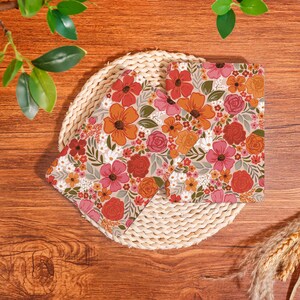 May include: Two floral print napkins with a beige background, featuring orange, red, and pink flowers, and green leaves. The napkins are placed on a round woven placemat, set against a wooden background.