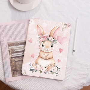 May include: A pink and white digital tablet case with a cute illustration of a brown bunny wearing a flower crown. The bunny is surrounded by pink hearts and flowers.