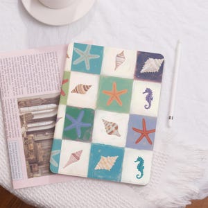 Pode incluir: Um caderno quadrado estampado com uma grelha de quadrados coloridos com ilustrações de conchas, estrelas do mar e cavalos-marinhos. O caderno tem um fundo branco e um estilo suave e artístico. Uma caneta branca está ao lado do caderno.