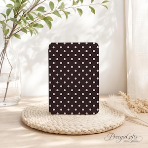 May include: A rectangular e-reader cover featuring a dark brown background with a repeating pattern of small, light pink polka dots. The cover is positioned upright on a woven circular mat, displaying a consistent grid of dots across its surface.