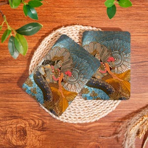 May include: Two rectangular coasters featuring a detailed illustration of a deity with dark skin, wearing gold jewelry and a yellow garment, holding a pink lotus flower. The background displays a blue mandala pattern and a multi-headed serpent.