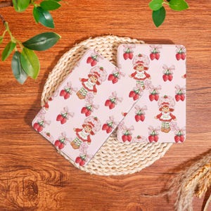 Puede incluir: Dos estuches rectangulares rosas con un patrón repetido de una niña de dibujos animados, fresas y ositos de peluche. Los estuches están sobre un salvamanteles tejido, con hojas verdes y espigas de trigo en el fondo. Los estuches son probablemente para guardar cosas.