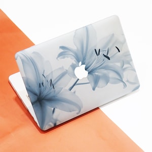 May include: A laptop with a floral design. The cover features a large, light blue lily flower print against a white background. The Apple logo is visible. The laptop is open, revealing the keyboard and screen.