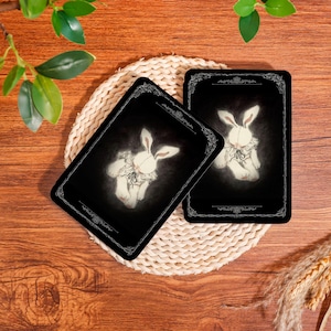 Puede incluir: Dos tarjetas rectangulares negras con una ilustración de un conejo blanco. El conejo lleva un cuello con volantes y un lazo. Las tarjetas tienen un borde decorativo y están sobre una esterilla tejida, con un fondo de madera.