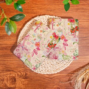 May include: Two floral-patterned notebooks with a beige background, featuring illustrations of flowers, tomatoes, and bows. The notebooks are placed on a woven mat, with text that reads "I'm so proud of you."