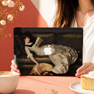 Peut inclure: Un autocollant pour ordinateur portable représentant une peinture d'une personne en robe blanche allongée sur un canapé sombre, lisant un livre et caressant un chien. Le logo Apple blanc est visible au centre de l'image sur l'ordinateur portable.