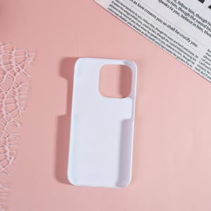 May include: A white phone case with cutouts for the camera and buttons. The case is made of a smooth material and is designed to fit a smartphone. The background is a light pink color with a piece of paper with text.