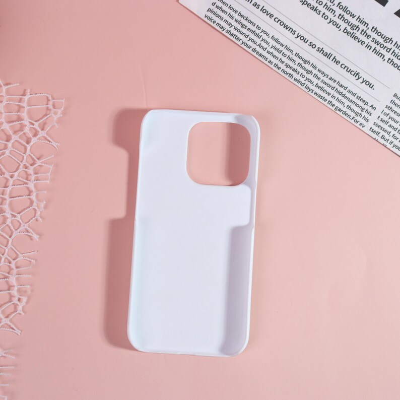 May include: A white phone case with cutouts for the camera and buttons. The case is made of a smooth material and is designed to fit a smartphone. The background is a light pink color with a piece of paper with text.