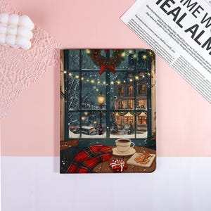 May include: A notebook with a winter scene illustration. The image depicts a cozy interior with a window overlooking a snowy street with buildings, a wreath, and string lights. A cup of coffee, cookies, and a gift box are on a table.