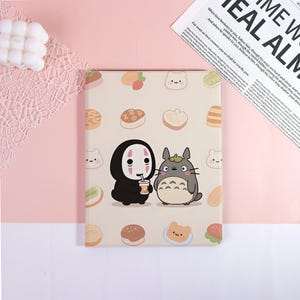 May include: A beige notebook featuring cartoon characters and food illustrations. The cover showcases a black figure with a white mask and a gray, furry creature. The notebook is decorated with various food icons.