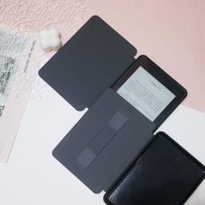 May include: A gray e-reader case with a black e-reader inside. The case has a built-in stand and a strap to hold the e-reader in place. The e-reader screen displays the text "Le Chapitre VI".