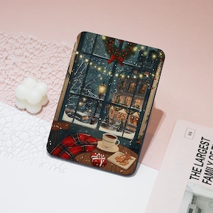 May include: A rectangular decorative item featuring a winter scene through a window. The illustration shows a cozy interior with a cup of coffee, gingerbread cookies, and a gift box. Outside, a snowy street with buildings and Christmas lights is visible.