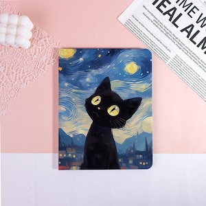 May include: A notebook with a black cat design set against a starry night sky, in the style of Van Gogh. The cat has yellow eyes and is set against a town silhouette. The notebook is on a pink surface.