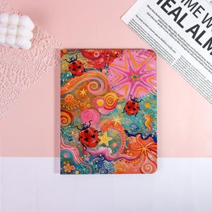 May include: A rectangular notebook cover featuring a vibrant, multi-colored illustration of three red ladybugs with black heart-shaped spots. The background is filled with swirling patterns, yellow stars, orange sea creatures, and pink floral shapes.
