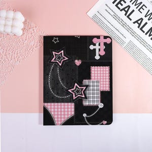 May include: Black notebook with pink and white star and heart designs. Features a patchwork pattern with gingham and zipper details. Includes a chain and a cross design. The notebook is on a pink surface.