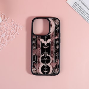 May include: Black phone case with a decorative design featuring bats, moons, and floral elements. The case has a dark background with a pink and white marbled effect. The case has a black border.