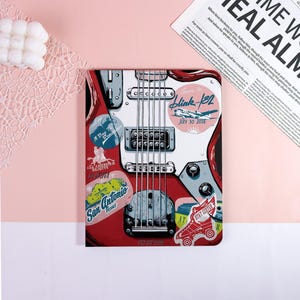 May include: A red and white guitar-shaped notebook with stickers. The stickers include text that says "San Antonio Texas", "Blink-182 July 30 2016", and "A Day To Remember". The notebook is on a pink surface.